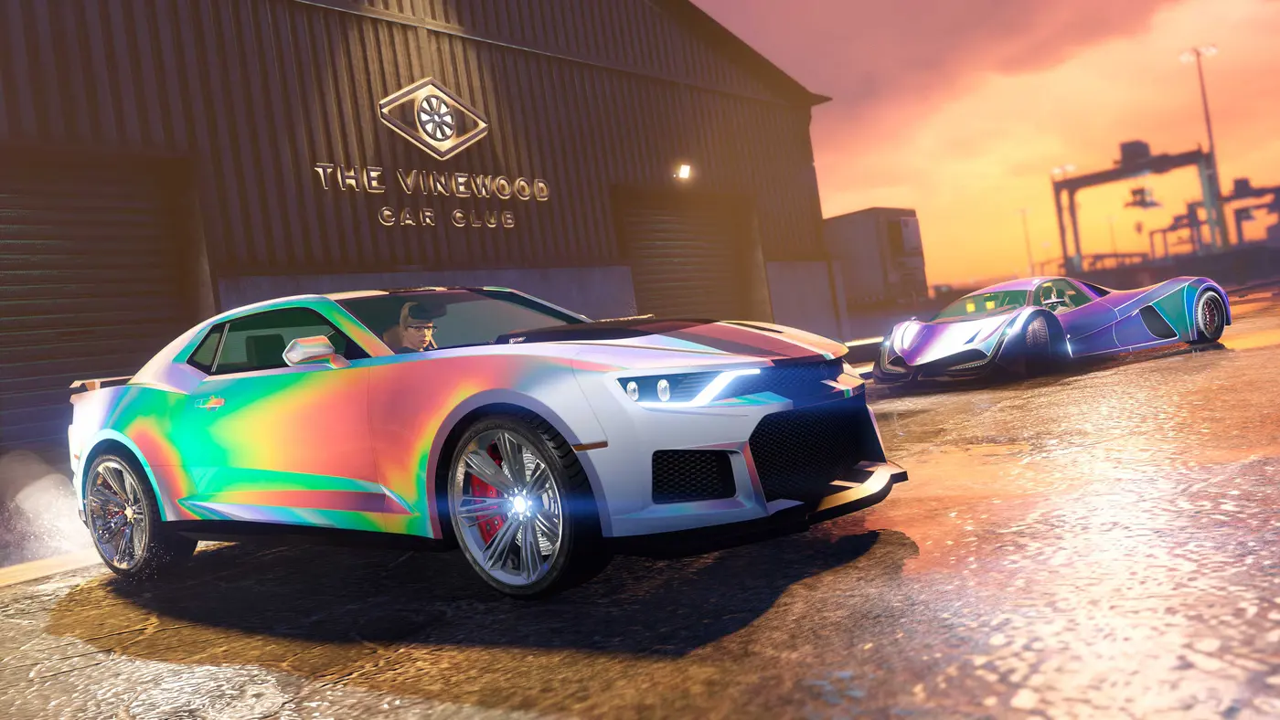 GTA Online screenshot: two vehicles with iridescent chameleon paint parked outside the Vinewood Car Club warehouse at sunset on Elysian Island, harbor cranes in the background